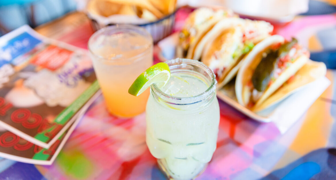 Margarita in a mason jar garnished with a lime, as well as three tacos line up on a table.