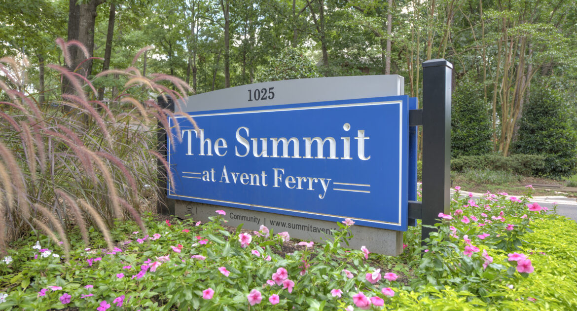 Outdoor signage of "The Summit at Avent Ferry". The sign is blue and being displayed in a bright green and pink flowered garden.