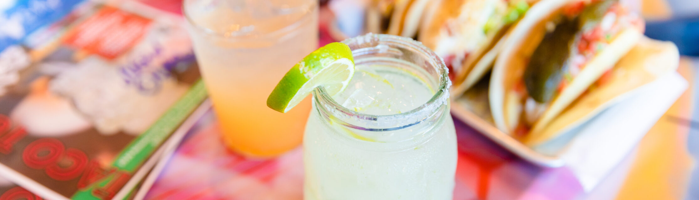 Margarita in a mason jar garnished with a lime, as well as three tacos line up on a table.